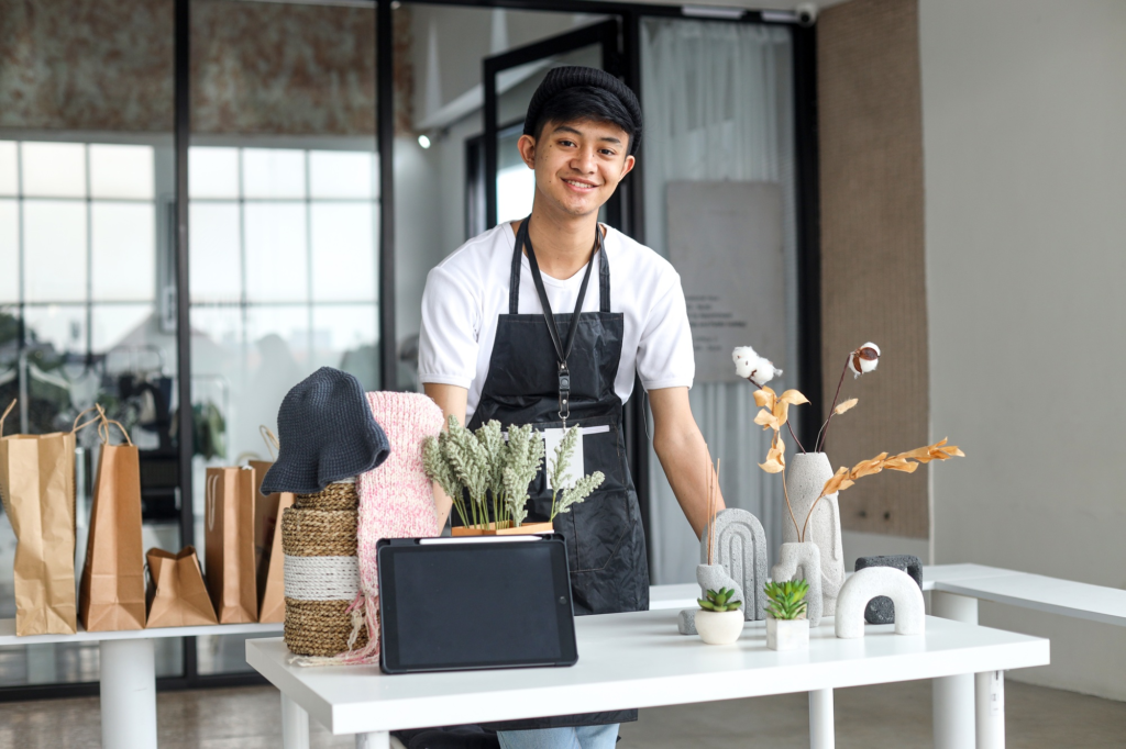 A smiling young artisan at a minimalist home decor booth showcasing handmade concrete vases, dried floral arrangements, and small potted plants.